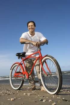 Caucasian mid-adult man posing with red bicycle on beach.