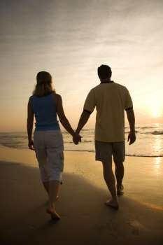 Caucasian mid-adult couple walking holding hands on beach at sunset.