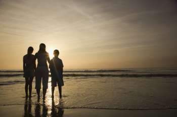Caucasian mid-adult mother and teenage kids standing silhouetted on beach at sunset.