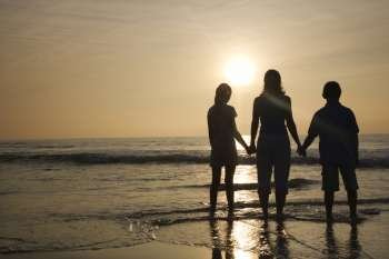 Caucasian mid-adult mother and tenage kids standing silhouetted on beach at sunset.
