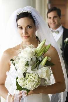 Caucasian mid-adult bride holding bouquet with groom in background.