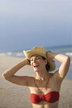 Caucasian mid-adult female laughing on beach in swimsuit holding hat on head.