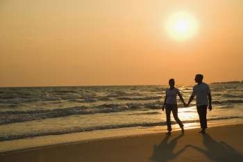 Caucasian mid-adult couple holding hands and walking down beach at sunset.