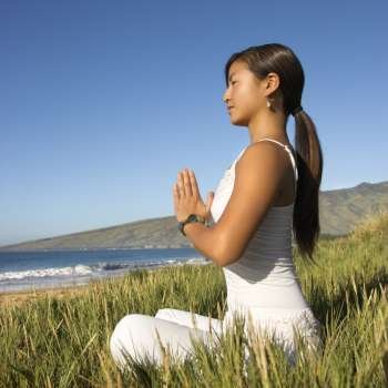 Young Asian female sitting on beach looking out to ocean with hands pressed together.