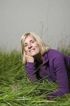 Middle-aged Caucasian woman lying in grass resting head on hand smiling.