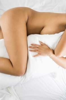 Nude woman hugging pillow lying on bed with white sheets.