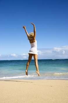 Pretty young woman jumping on Maui, Hawaii beach with arms raised into air.