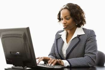 Businesswoman sitting at office desk typing on computer and talking into telephone headset.