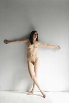 Nude woman against wall with legs crossed and arms out to side.