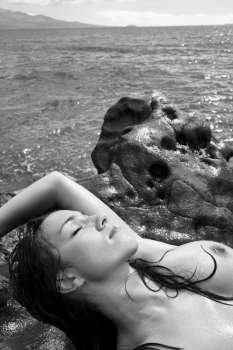 Topless Caucasian mid adult woman on rocky Maui coast lying on back with eyes closed.