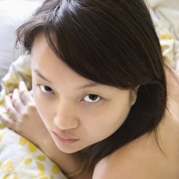 Head and shoulder portrait of pretty young Asian woman lying in bed making eye contact.
