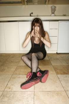 Pretty Caucasian young woman sitting on kitchen floor holding hands over mouth.