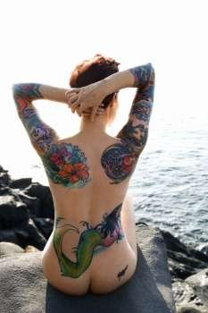 Sexy nude tattooed Caucasian woman sitting on rock on beach in Maui, Hawaii, USA.
