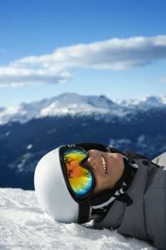 Caucasian teenage boy snowboarder wearing helmet and goggles, smiling, looking at viewer, lying in snow on mountain with mountain landscape in background.