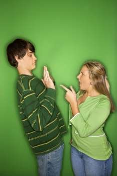 Portrait of Caucasian teen girl pointing at yelling at teen boy who is holding up hands to her against green background.