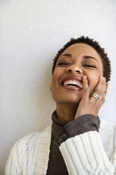 Close up head and shoulder of African-American woman standing against white wall smiling with hand on face and eyes closed.