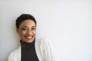 Close up of African- American woman standing against white wall looking at viewer and smiling.