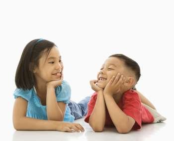 Young Asian brother and sister lying on floor with head on hands looking at eachother smiling.
