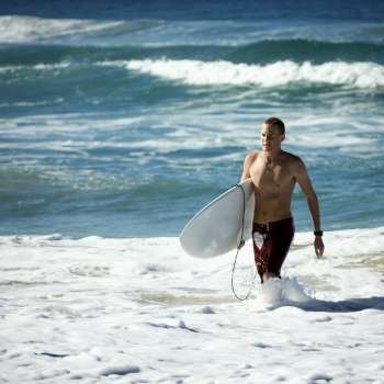 Young adult male walking ocean carrying surfboard in Surfers Paradise, Australia.