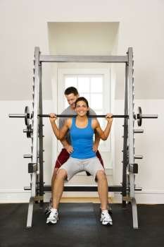 Woman lifting weights in gym being assisted by man.