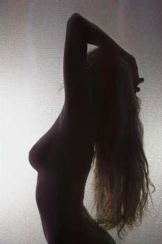 Side view of nude Caucasian woman silhouetted behind sheer cloth with hand to head and long hair.