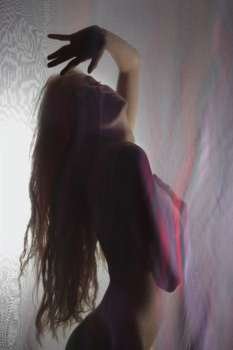 Nude Caucasian woman backlit behind sheer cloth with hand raised and long hair.