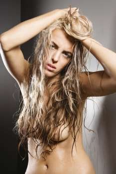 Nude Caucasian woman with hands in long blond hair looking at viewer.