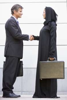 Two businesspeople standing outdoors with briefcases shaking hands and smiling
