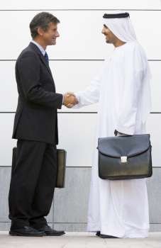Two businessmen standing outdoors with briefcases shaking hands and smiling