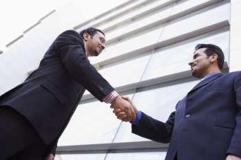 Two businessmen outdoors by building shaking hands and smiling (high key/selective focus)