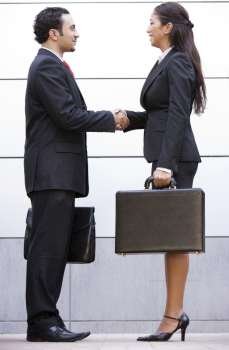 Two businesspeople shaking hands outdoors holding briefcases and smiling