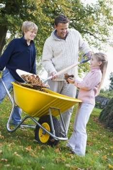 Father outdoors with two young children shoveling leaves into a wheelbarrow and smiling (selective focus)