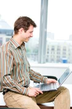 Happy young businessman sitting on table at office and using laptop computer, smilng.