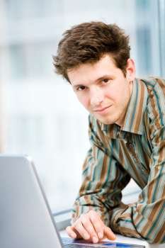Casual looking happy businessman working on laptop computer in front of office window, smiling.