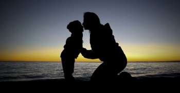 Mother kneels kissing child at waters edge, sunset