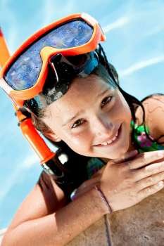 A happy young girl smiling and relaxing on the side of a swimming pool wearing orange goggles and snorkel
