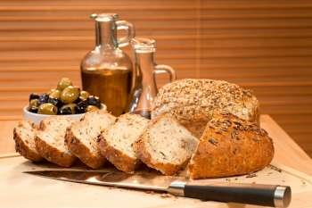 Freshly cut and sliced rustic seeded bread with a knife and a bowl of green and black italian olives, a bottle of olive oil and balsamic vinegar. The focus is on the bread in the foreground.