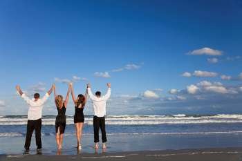 Four young people, two couples, holding hands, arms raised having fun and celebrating on a beach