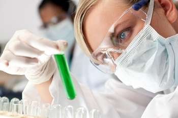 A female medical or scientific researcher or doctor looking at a green solution in a laboratory with her Asian female colleague out of focus behind her.