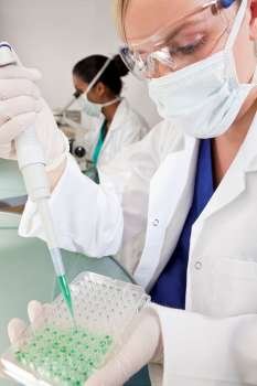 Environmental concept shot of a female medical or scientific researcher or doctor using a pipette and looking at a sample tray of a green solution in a laboratory with her Asian female colleague out of focus behind her.