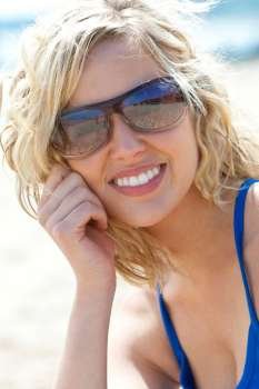 Close up portrait of a beautiful young blond woman with perfect teeth wearing sunglasses and smiling while sitting on a beach