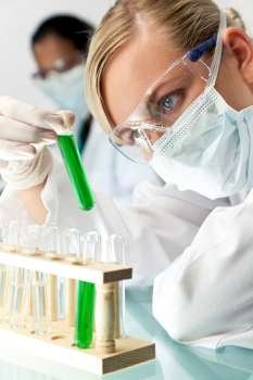 A blond medical or scientific researcher or doctor using looking at a test tube of green solution in a laboratory with her Asian female colleague out of focus behind her.
