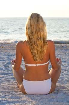 A beautiful young blond woman in a white bikini sits crosslegged on a beach at sunset