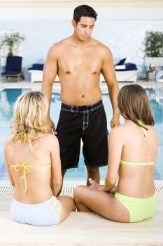 Mid adult man standing in front of two young women at the poolside