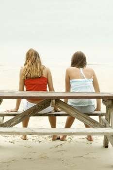 Rear view of girl and a teenage girl sitting on a picnic table on the beach