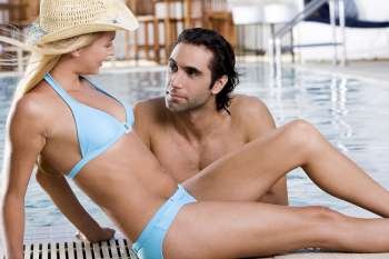 Side profile of a young woman sitting at the poolside with a mid adult man in the swimming pool