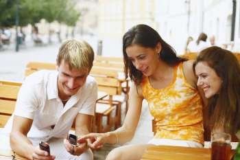 Young man holding two mobile phones with two young women sitting beside him 