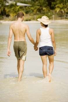 Rear view of a young man and a teenage girl holding hands walking on the beach