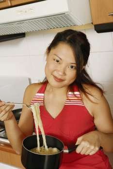Portrait of a young woman holding noodles with a fork at a kitchen counter