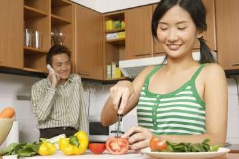 Close-up of a young woman cutting vegetables in the kitchen with a young man talking on a mobile phone